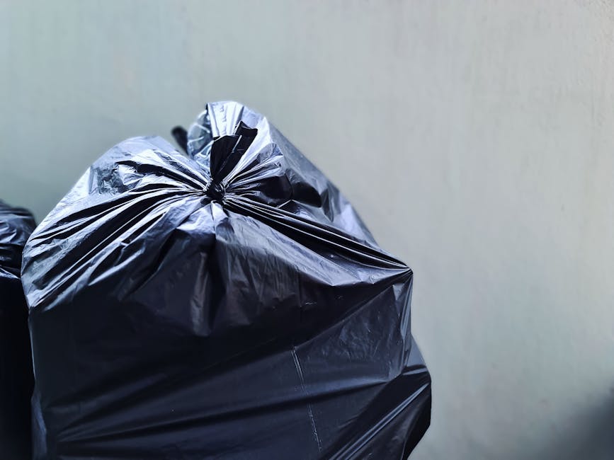A person wearing a white T-shirt and transparent disposable gloves is holding a white plastic rubbish bin labeled 'PLASTIC' on the front. The lid of the bin is open, revealing a black plastic bin line