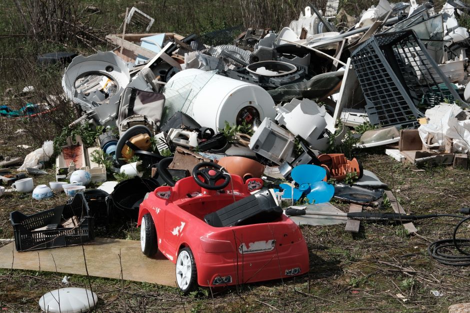 A large pile of discarded household and electronic waste sprawled across an outdoor area, consisting of various broken appliances, plastic items, and metal debris. Prominently visible in the foregroun